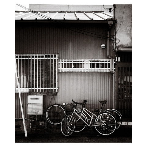 The photograph is a monochrome image of a residential area with a pair of bicycles equipped with front baskets and two spare tires beside them. Window bars and a bamboo curtain suggest measures against burglary. A gas water heater is installed outside, and small details like an aerosol can are visible through a narrow frosted glass window. An exhaust vent indicates the presence of appliances, alluding to a cautious yet comfortable home environment.