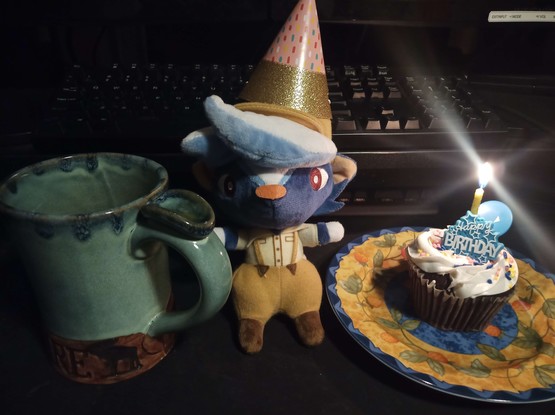 A plushie of a blue skunk named Kicks from animal crossing. They are looking directly at the viewer and wearing a small party hat. To the left of him is a coffee in a large mug, and to the right of him is a plated chocolate cupcake with a lit candle. There is a small sign on the cupcake which reads "happy birthday."