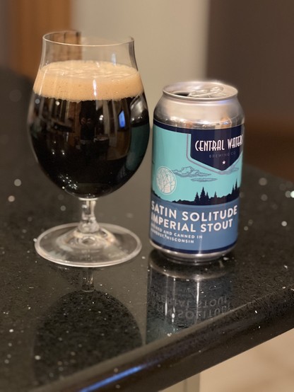 A tulip glass of Central Waters Satin Solitude imperial stout. The can is silver with an aquamarine blue label, white lettering, and a scene of trees and lakes.