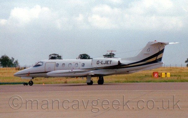 Side view of a white, twin engined bizjet with black and yellow cheatline running along the body, being towed from right to left, with grass and a handful of trees in the background, under a pale blue sky with bits of white fluff.