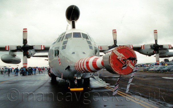 Almost head on front view of a grey, high-winged 4 propellor-engined military transport aircraft with a large black radar dome above the fuselage, and a long red and white probe extending forward from the nose, with a large cover on the end, under a grey sky.