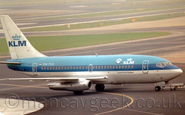 Side view of a blue and white, twin engined jet airliner with white "KLM" titles on the upper forward fuselage, and similar larger blue titles on the tail, being pushed back from its stand, from right to left, in front of a network of grass areas and taxiways stretching off into the distance, slowly vanishing in to haze.