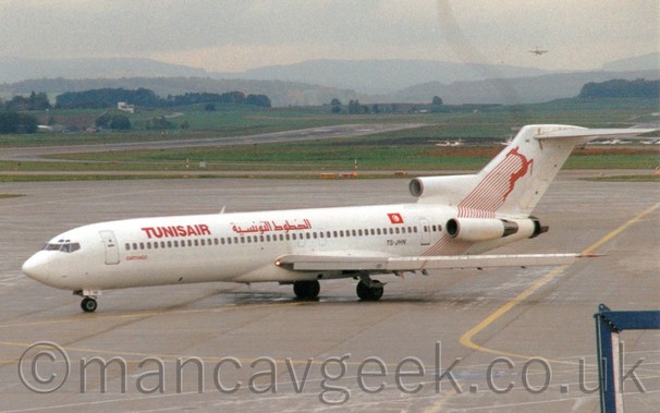 Side view of a white, 3 engined jet airliner with the engines mounted on the rear fuselage and red "TunisAir" titles in English and Arabic on the upper forward fuselage, and an image of a red leaping gazelle on the tail, taxiing from right to left, with grass in the background leading up to rolling hills in the distance. Another plane can be seen on approach to one of the runways.