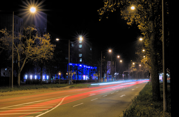 Foto einer Straße schräg von der Seite. Beide Seiten sind mit Laternen gesäumt, die sternförmige Halos bilden. Auf der Straße sieht man rote und weiße Lichtspuren. Dahinter ist ein blau angeleuchtetes Gebäude.