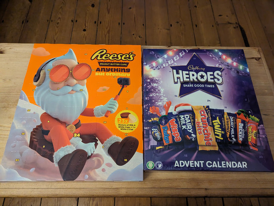 Two advent calendars sitting on a wooden table with the wooden flooring of the room it's in behind. The calendar on the left is a Reese's Peanut Butter Cups one; orange in the main, with a Santa in orange sliding down a snowy mountain on  peanut butter cup, holding a camera on a selfie stick. The other calendar is a Cadbury Heroes one; purple coloured with pictures of all the chocolates listed inside: Eclairs, Fudge, Wispa, Dairy Milk, Crunchie Bits, Twirl, Caramel, Double Decker, and Twisted.