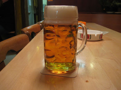 A half litre of Lager sitting on a pine-topped table.