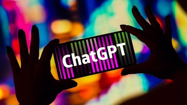 A brightly coloured background showing out-of-focus blobs of all colours. In the foreground, two silhouetted hands are holding a phone, with a screen displaying the word "ChatGPT"