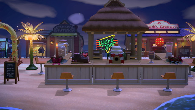 Animal Crossing: New Horizons screenshot. Image intended to look like a beachfront bar with white barfront / wooden tabletops and stools set up. On the two ends of the bar are holiday candles. There are various drinks on the counter and a yule log. There is a beachy canopy structure in the middle for the bartender to stand under. On either side of the bar near the back are palm tree lamps. Visible behind the bar are the Able Sisters shop and Nook's Cranny.