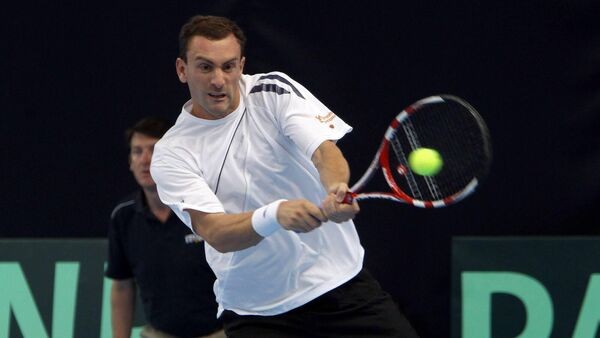 A photograph of Irish tennis player Conor Niland. He is seen mid-shot, in a white t-shirt and black shorts. He is hitting a tennis ball on his left-hand side with a back-handed shot. Behind him is a ball boy watching on