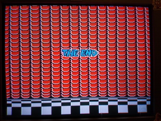 Screenshot of the final screen of Super Mario bros 3. Shows a red curtain descended to a black and white checkered floor with the words "The End" in blue letters at the center of the screen.