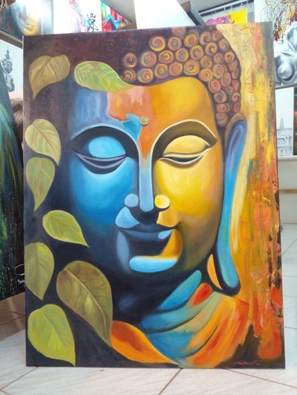 a painting of the Buddha, from his collarbones up, half his face is blue and the other half orange, the left side of the painting is shadowy, with green leaves coming from the dark