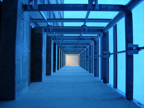 Color photo looking directly up through an open shaft that is defined by one wall of balconies (left), one smooth concrete wall (bottom), and two walls of metal framed blue glass (right and top)