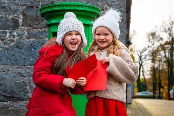 A photograph of two little girls dressed up in warm winter clothing laughing while looking at the camera. They are each holding red envelopes and are stood next to a bright green post box