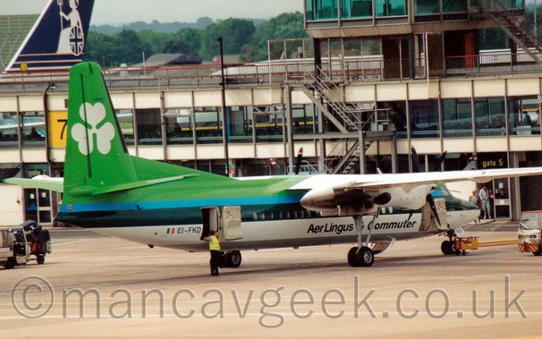 Side view of a green and white, high-winged, twin propellor-engined airliner parked facing to the right, and slightly away from the camera, in front of a terminal building with a short, highly glazed metal-framed tower.