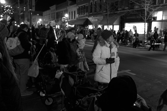 A busy picture of people watching a parade. Both sides of the street are lined with people. OF the many only one person is a standout. She is wearing a white coat, everyone else is dressed very dark