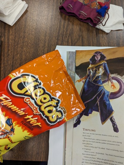 Bag of Flamin' Hot Cheetos next to an image of a Tiefling from the DnD 5e Player Guide. The Tiefling has purple skin, but the hands are glowing orange.