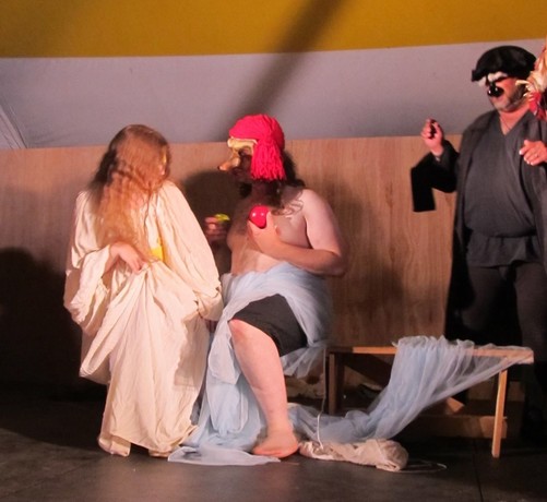 Three actors perform a commedia dell'arte play on a raised wooden stage in a pavilion tent during a medieval camping event portraying the stock characters of Isabella (only in a paint smeared chemise) flirting with il Capitano (disguised as a nearly nude Isabella for a painting) on a bench while being observed by il Dotore (and also Pantalone just off frame).