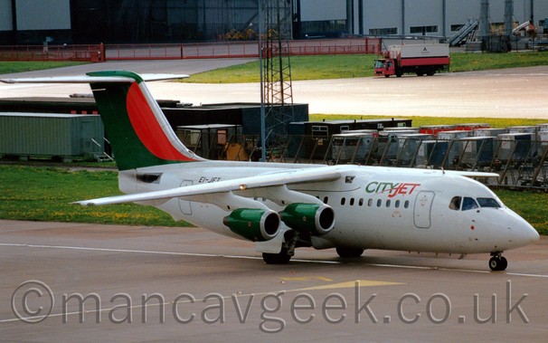 Side view of a white, high-winged, 4 engined jet airliner, with green and red "CityJet" titles on the upper forward fuselage, green engine pods, and a green and red tail, taxiing from left to right in front of some low buildings and luggage containers, with a large grey hangar in the distance.