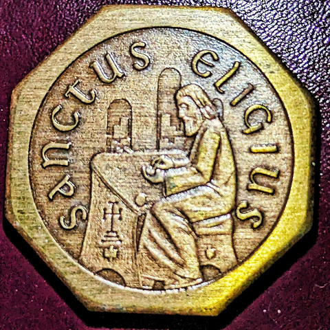 An Octagonal medal with all details in a recessed circule. "Sanctus Eligius" (Saint Eligius, in Latin) around the edge, the saint himself sitting at a desk looking at coins. The desk has a representation of the Cross atop Mount Calvary similar to that depicted on Byzantine coins of the time of Saint Eligius.