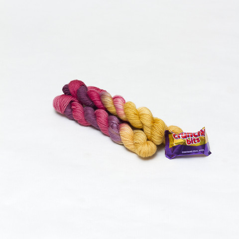 Picture shows three mini skeins of yarn positioned diagonally in a pyramid. The yarn is red, purple and gold. Next to the yarn is a small foil wrapped Crunchie chocolate.