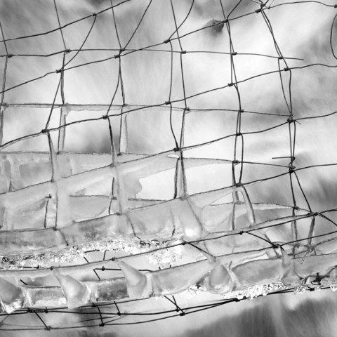 A black and white long exposure photography of a wire fence over a stream partially encased in ice