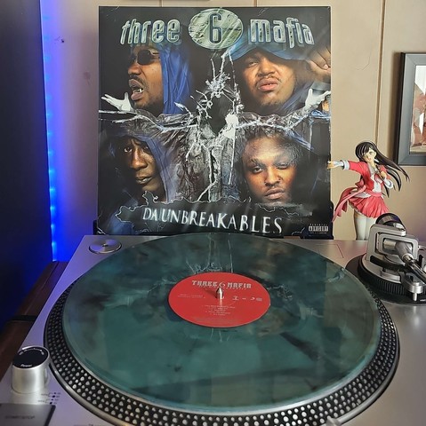 A Translucent Marbled Cloudy vinyl record sits on a turntable. Behind the turntable, a vinyl album outer sleeve is displayed. The front cover shows 4 members of Three 6 Mafia and a black masked figure with its arms out.