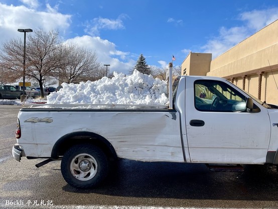 A truck parked near a building. Thereâ€™s a huge pile of snow in its trunk.