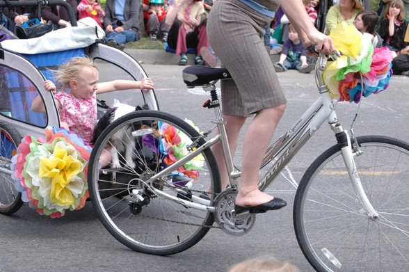A little kid with windblown hair rises up in a bike trailer, with a dog as a person pedals a bike in front. A large tissue paper flower is affixed to the handle bares and also one on the trailer.