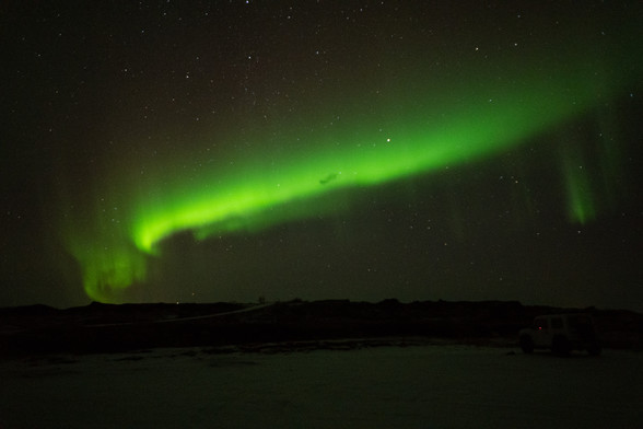 Green aurora borealis (northern lights) above a dark landscape, with some snow visible. There is a car at the bottom left