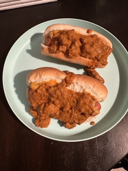 The picture shows two chili hot dogs on a plate. The hot dogs are in soft buns and are generously topped with a chunky chili sauce. The chili sauce appears to be tomato-based with ground meat. The plate is round and has a light blue color. The plate is placed on a dark brown table, and there is a white napkin or cloth visible in the top left corner of the picture. The chili on the hot dogs looks rich and hearty, and some of it is spilling onto the plate.