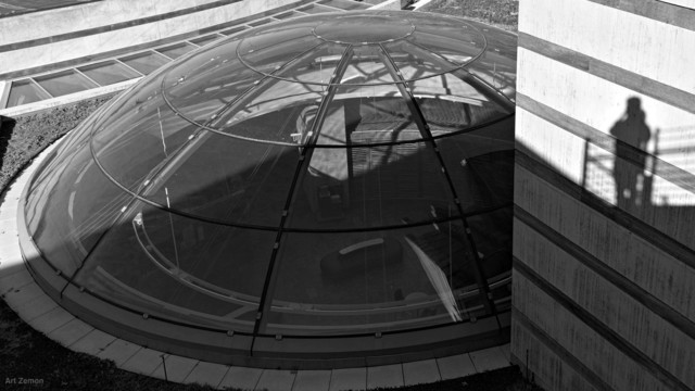 black and white photo looking down on the glass dome of the Crystal Bridges Museum of American Art. The shadow of the photographer appears on the right