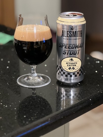 A silver pint beer can has black lettering and yellow accents. A yellow oval badge states “Affogato Edition” and “Sprcual Edition” with Ale Smith’s trademark checkered flags crossed in the middle.
A tulip cup of dark beer has a smooth tan creamy head.