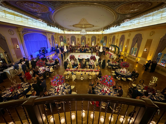 The historic Palais Royale Ballroom in South Bend IN. Seen from the balcony, it is beautifully decorated for a wedding with round tables everywhere, accents of red and green and a warm golden Hugh to the entire room. Beautiful artwork on the ceiling with a large chandelier, and the band stage lit with blue lights on the left.