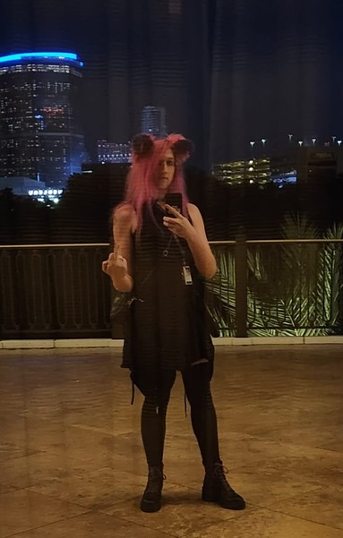 My reflection, on a balcony at the Wynn, Vegas  skyline behind me.  I'm wearing a black dress, pink+purple cat ears to match my hair, and a hoodie around my waste.  Black boots w/ colorful laces & thigh high socks.  Leather-ish purse.  Conference Badge around my neck.  I'm flipping off the camera.