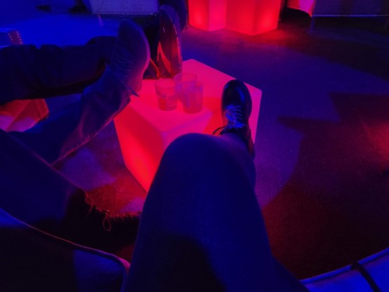 Some people resting their feet on an red, illuminated cube.  Blue/red/purple/bisexual lighting.  Some empty drinks.  At re:Play.
