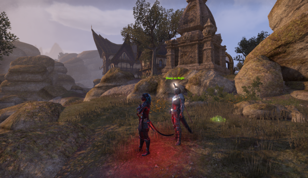 In-game screenshot (with UI hidden) of the outskirts of Rawl'kha in Reaper's March, with a few Khajiiti style buildings cresting a rocky arid ridge with more rocky outcroppings on the horizon. In the foreground stand my Khajiit toon, my Argonian companion, and my voriplasm pet; my toon is suffused with a red glow from one of his equipped skills.