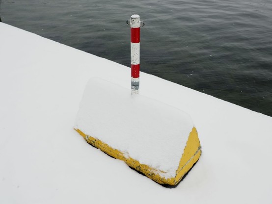 picture of a red-white pole and a yellow concrete block in the snow. Water in the background