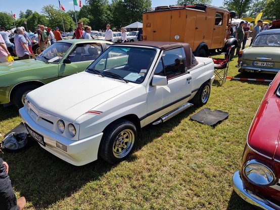 A small white hatchback with a folding roof. It is parked in a field surrounded by other classic cars. It has some aftermarket components, such as a body kit, alloy wheels, and a side exhaust. Hmm no I'm posting picture to mastodon but using voice dictation.