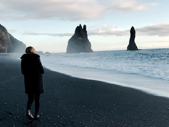Photo of a woman looking across the water at two large rock formations while walking on a beach in Iceland