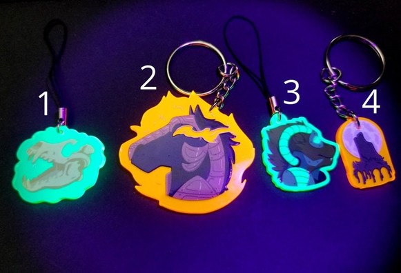 Photo of 4 blacklight reactive (glows) keychains/charms. Far left is a canijr skull with open maw and whisks of magic or ghostly presence on green acrylic. Next is an armored horse bust, "nightmare" with glowing eyes on orange acrylic. Next is a cyborg critter with glowing eyes and glowing tunes on green acrylic. Far right is a howling wolf bust in front of a moon on orange acrylic.