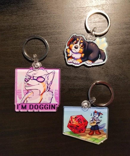 Photo of 3 keychains, top is a bernese mountain dog pup on a blue background, middle is a vaporwave styles Australian shepherd anthro character with sunglasses on a grid background with blue palm trees and says "I'm doggin" in a pixel font. Bottom is a Tabletop game grid with an chibi/small miniature of a wolf anthro in a bard outfit playing a lute with a normal sized D20/dice next to him on a blue background.