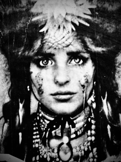 A monochrome Hipstamatic photo of a piece of street art showing the head of a young woman wearing an elaborate headdress and many strings of beads