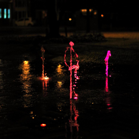 Photograph of a fountain lighted from beneath; photo is taken at night against dark background. Water in the fountain is a single stream up; the water coincidentally takes the shape of a head, two arms and legs, with one leg up as if it is a dancing stick figure