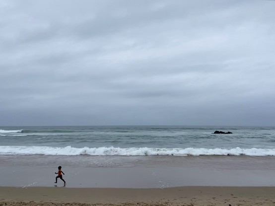 View on the side of a child running on an empty beach right by the water; gloomy day; horizon a straight line; mostly blue and grey tones; some waves and foam