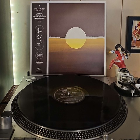A black vinyl record sits on a turntable. Behind the turntable, a vinyl album outer sleeve is displayed. The front cover shows a rising sunrise painting