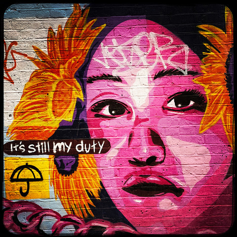 A colour Hipstamatic photo of a piece of street art showing a young woman's face, wearing sunflowers around her ears, with the words, "Still my duty" to her left