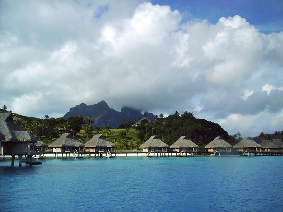 A picture taken while snorkeling in Moorea (in French Polynesia).  Huts on stilts are shown in the water near a beach with a mountain in the background.