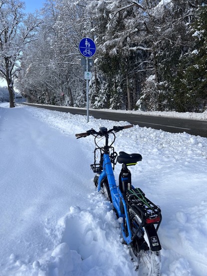 blaues kompaktrad steckt bis zur mitte der 20-zoll-räder im schnee. daneben ein schild zur gemeinsamen nutzung des weges von fußgehenden und radfahrenden. die bäume sind schneebedeckt.