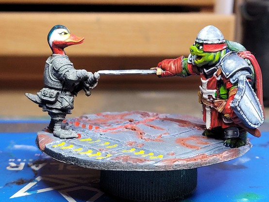 The previous diorama, but now the duck is standing opposite the turtle.  Only the duck's head is panted so far, with orange-red bill, a stripe of black running from front to back up the throat, nose, and over the head to the back, dark turquoise on top, yellow shading back into white around the eyes, running back down the head to a point, and red cheeks and throat between the stripe of black and the area of white near the eyes.

Overall, it looks roughly like a Mandarin duck.