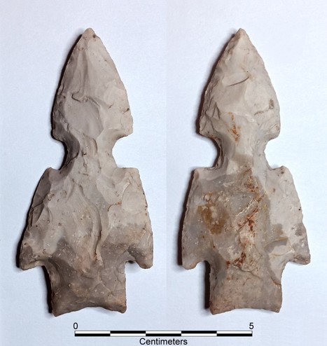 White & medium gray smooth chert artifacts. It has a concave base, straight stem, & strong, slightly barbed shoulders formed by basal notching. The long triangular blade is interrupted about halfway up with a set of deep, wide notches.